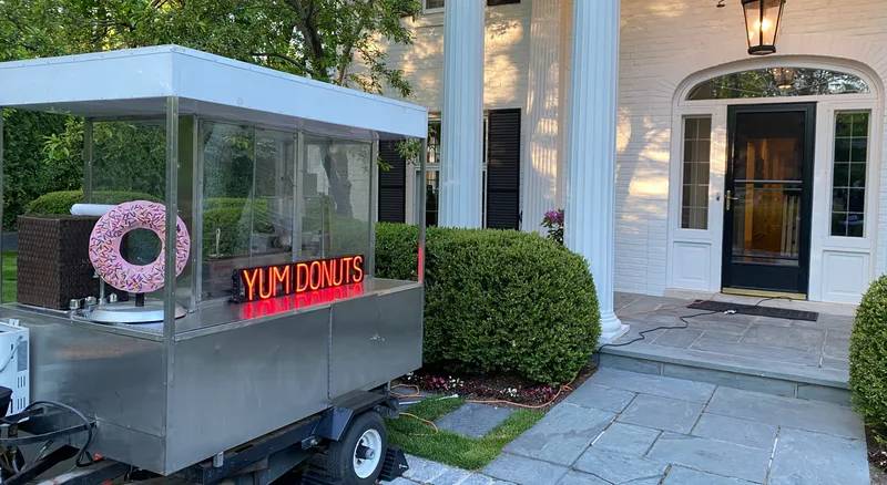 The Happy Dough Lucky mini donut trailer with its neon YUM DONUTS sign parked outside a house